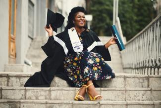 Woman in grad cap and gown sitting on stairs and smiling. She's wearing a ribbon that says Doctor of Medicine